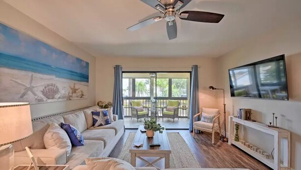 A spacious living room with a ceiling fan, a large painting of a beach scene, a sofa set, a coffee table, and a television on the wall.