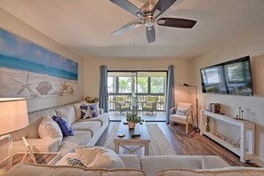 Un salon spacieux doté d’un ventilateur de plafond, d’un grand tableau représentant une scène de plage, d’un ensemble de canapés, d’une table basse et d’un téléviseur fixé au mur.
