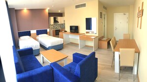 Habitación de hotel con dos camas, un escritorio, un televisor, una pequeña cocina y una zona de estar con sofás azules.
