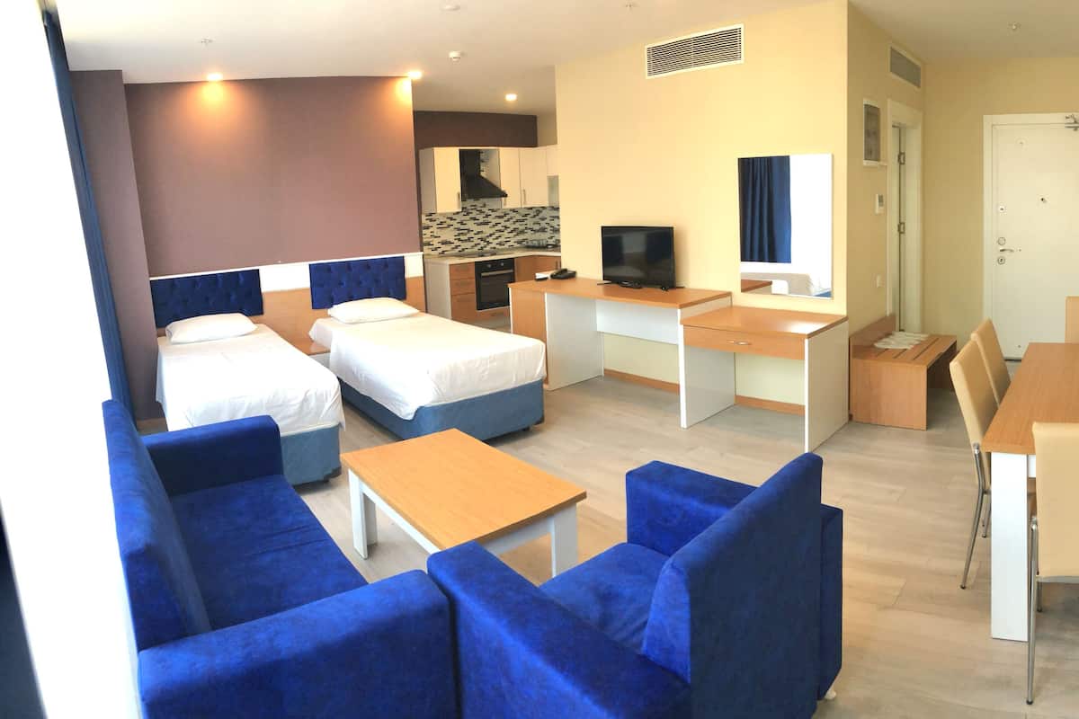 Une chambre d’hôtel avec deux lits, un bureau, une télévision, un petit coin cuisine et un espace salon avec des canapés bleus.
