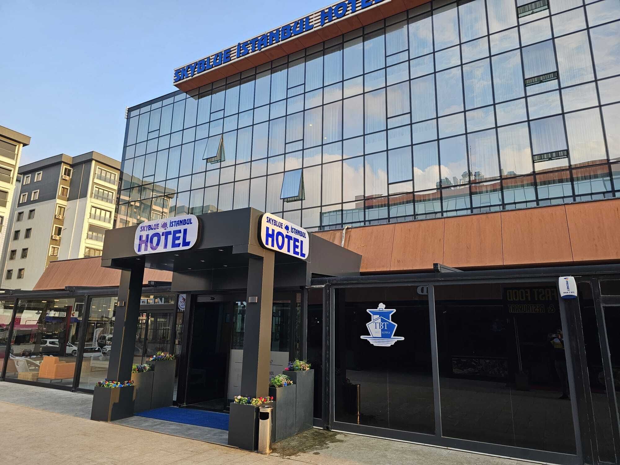 A modern hotel with a glass facade, two signs reading "Skyblue Istanbul Hotel", and a logo for "TBT".