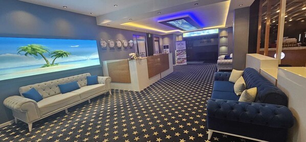 Un vestíbulo de hotel con un mostrador de recepción, dos sofás, una pantalla grande mostrando una escena de playa y una alfombra estampada.