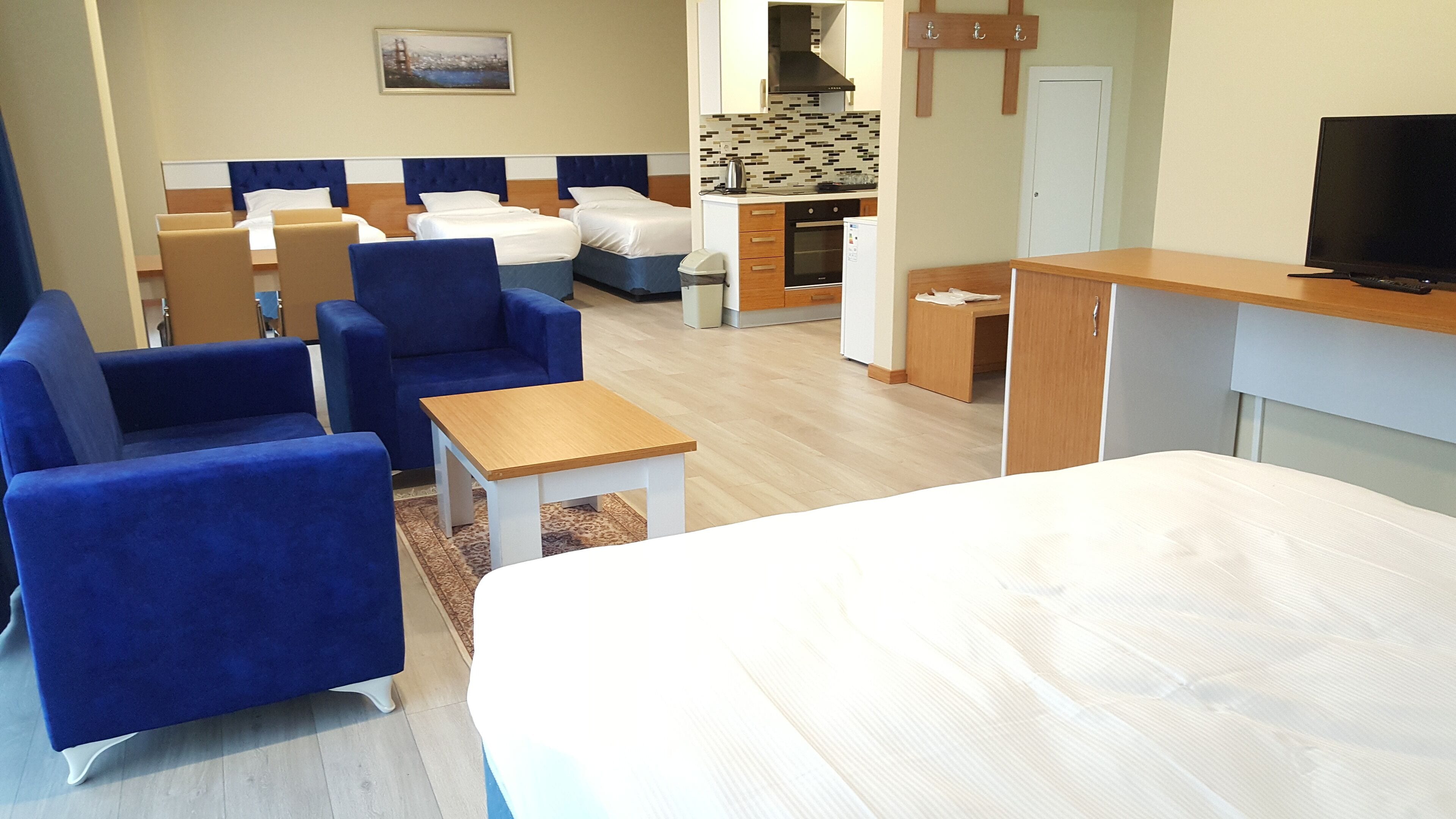 A hotel room with a bed, two blue armchairs, a small wooden table, a kitchenette, and a television.