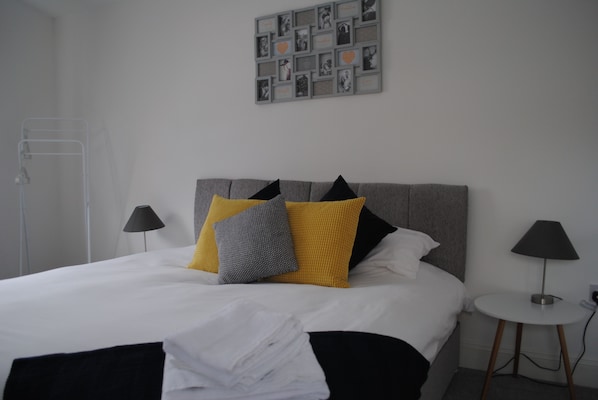 A neatly made bed with white linens and multiple pillows, a bedside table with a lamp, and a wall with framed photos.