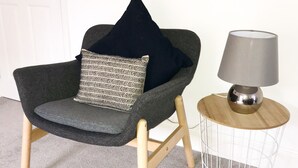 Een grijze fauteuil met een kussen met dessin, een klein rond tafeltje met een lamp, en een witte deur op de achtergrond.