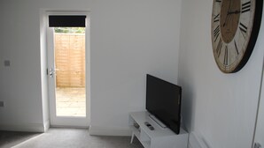 Een kamer met een witte deur die uitkomt op een houten terras, een flatscreen televisie op een witte standaard en een grote wandklok.