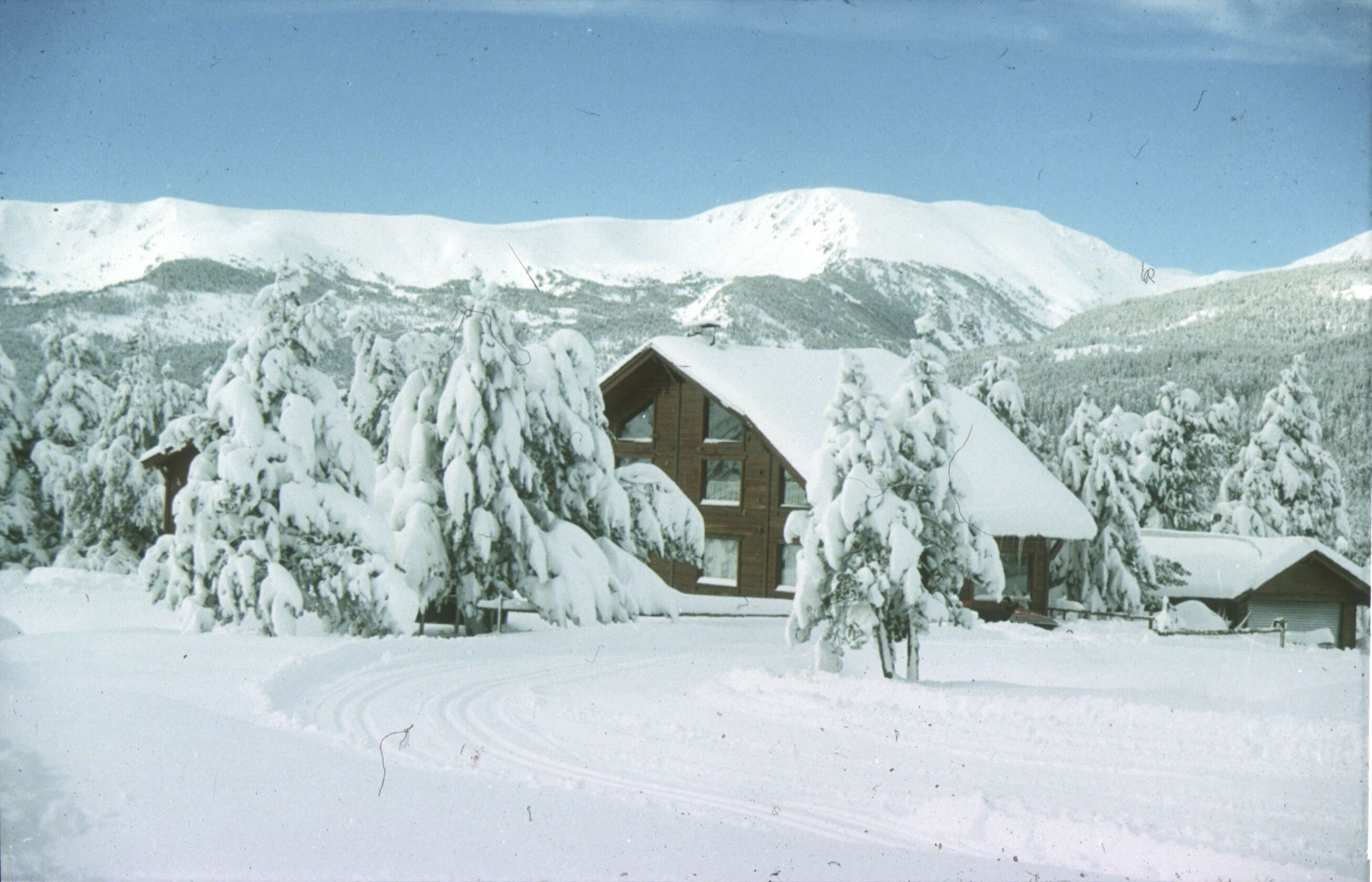 A snow-covered lodge with a wooden exterior, surrounded by snow-laden trees and mountains in the background.