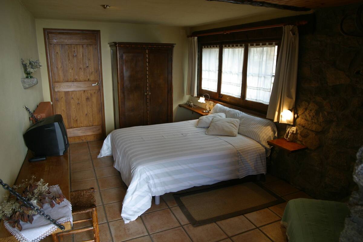 A bedroom with a large bed, a wooden wardrobe, a desk with a lamp, and a stone wall.