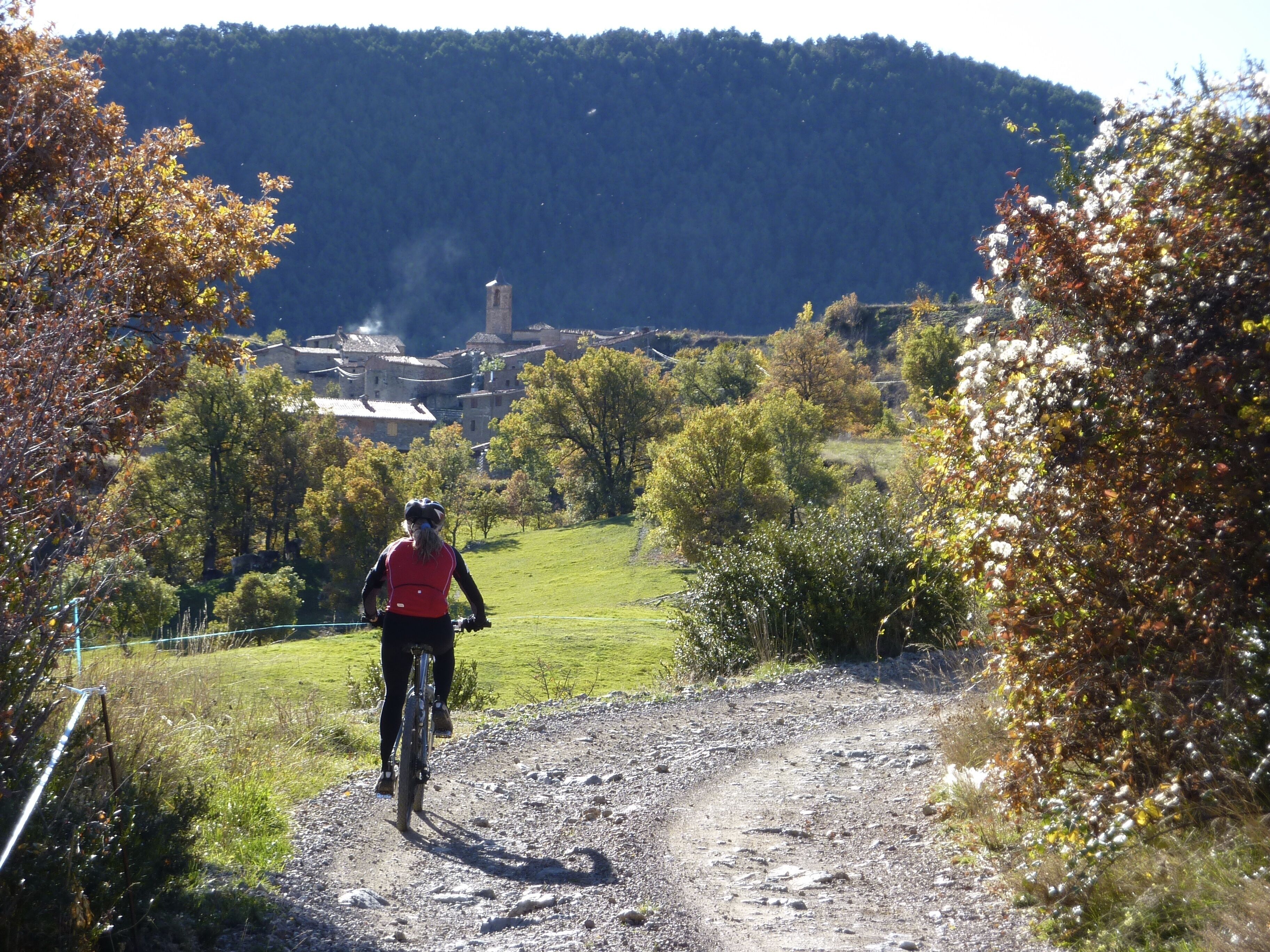 Um ciclista numa estrada de cascalho, com montanhas ao fundo e uma aldeia distante.