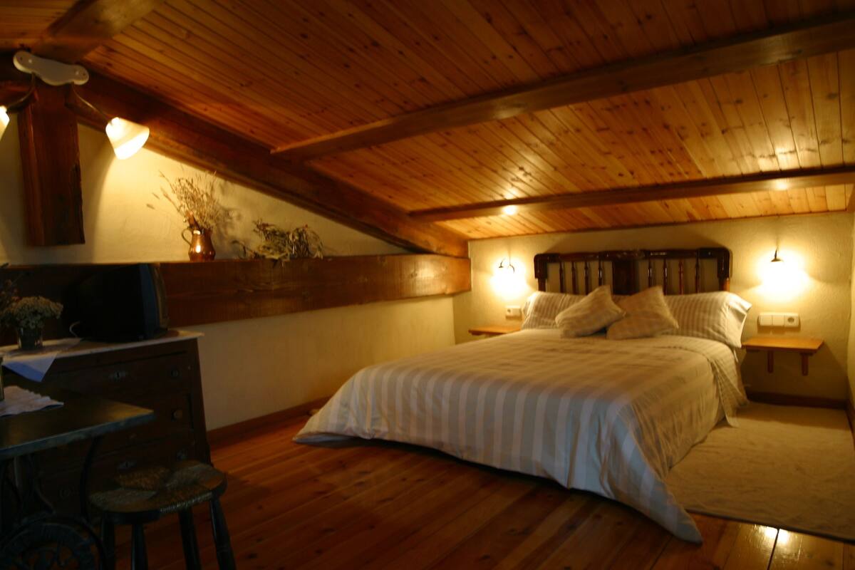 A bedroom with a wooden ceiling, a bed with a striped bedspread, a nightstand with a lamp, and a dresser with a vase of flowers.