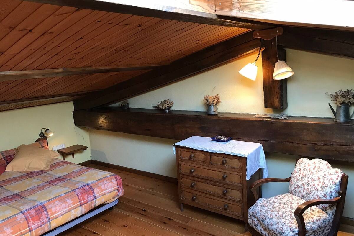 A cozy attic bedroom with a wooden ceiling, a bed with a plaid bedspread, a dresser, a chair, and a small table with a lamp.