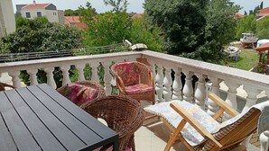 Un balcón con sillas de mimbre y una mesa, con vista a un jardín y edificios.