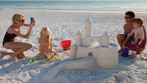 Een strandscène met een zandkasteel, een kind en een volwassene.