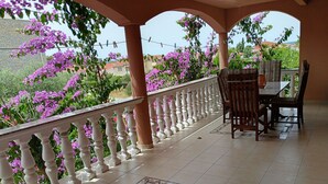 En balkon med hvid gelænder, et bord med stole, og lilla blomster i baggrunden.