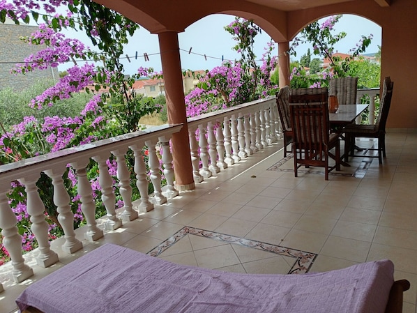 A balcony with a white balustrade, a table with chairs, and purple flowers in the background.