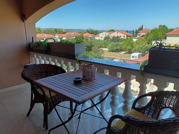 A balcony with wicker chairs, a wooden table, and a view of houses and mountains.