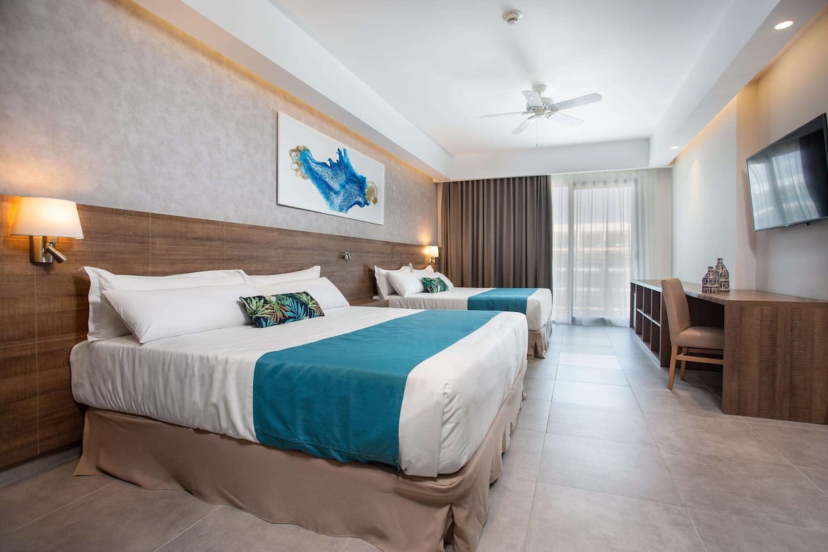 A hotel room with two beds, a desk, a TV, and a ceiling fan.