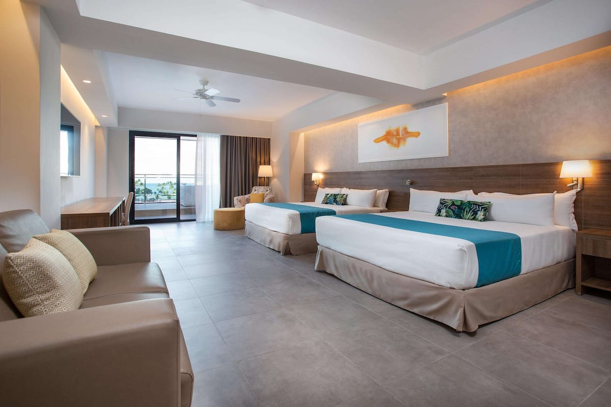 A modern hotel room with two beds, a sofa, a desk, and a balcony with a view.