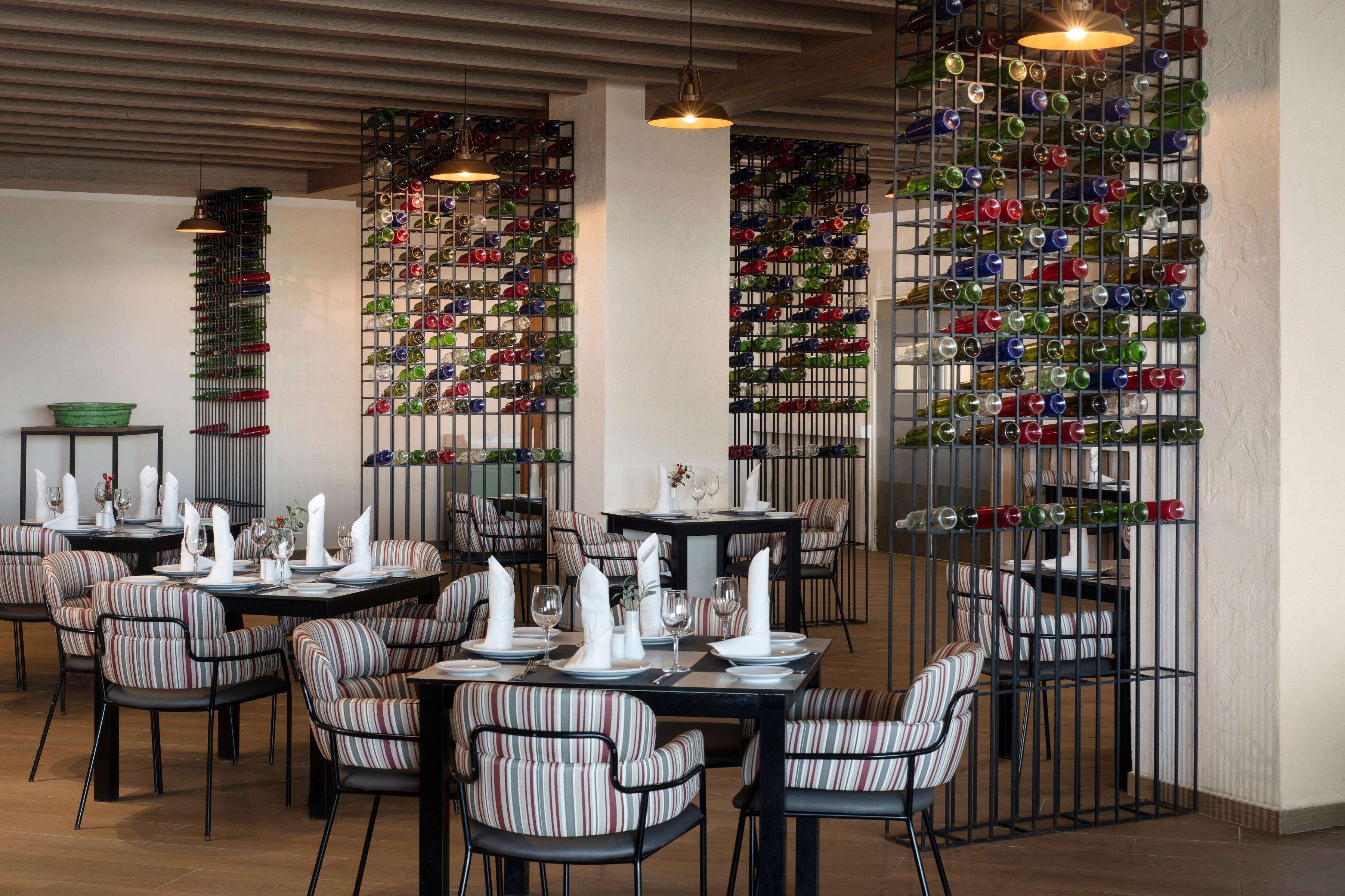 A restaurant interior with striped upholstered chairs, tables set with white napkins and plates, and a wall of colorful wine bottles.