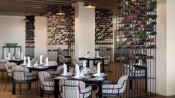 A restaurant interior with striped upholstered chairs, tables set with white napkins and plates, and a wall of colorful wine bottles.