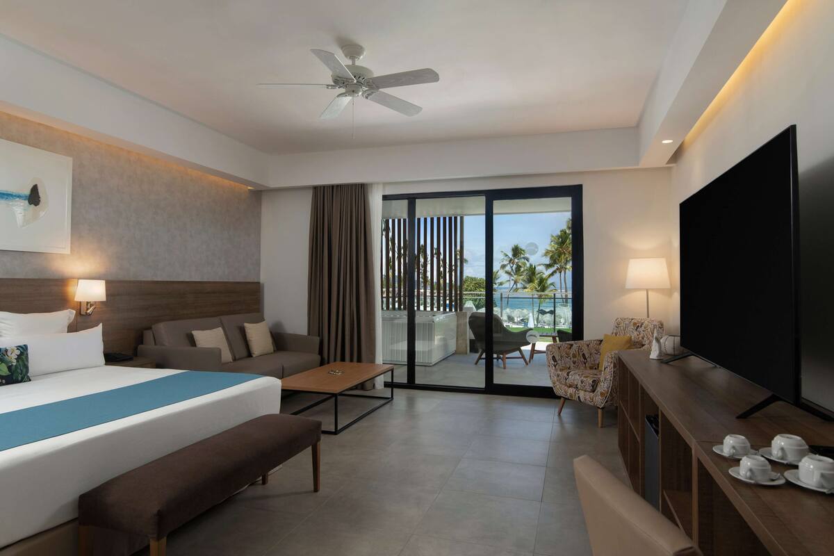 A modern hotel room with a large bed, a flat-screen TV, a ceiling fan, a sofa, a coffee table, and a balcony with a view of palm trees.