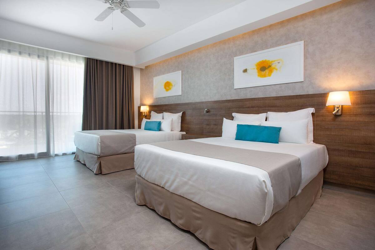A hotel room with two beds, a large window with curtains, a ceiling fan, and artwork on the walls.