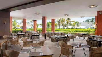 An outdoor dining area with wicker furniture and tables, surrounded by palm trees and a view of the ocean.