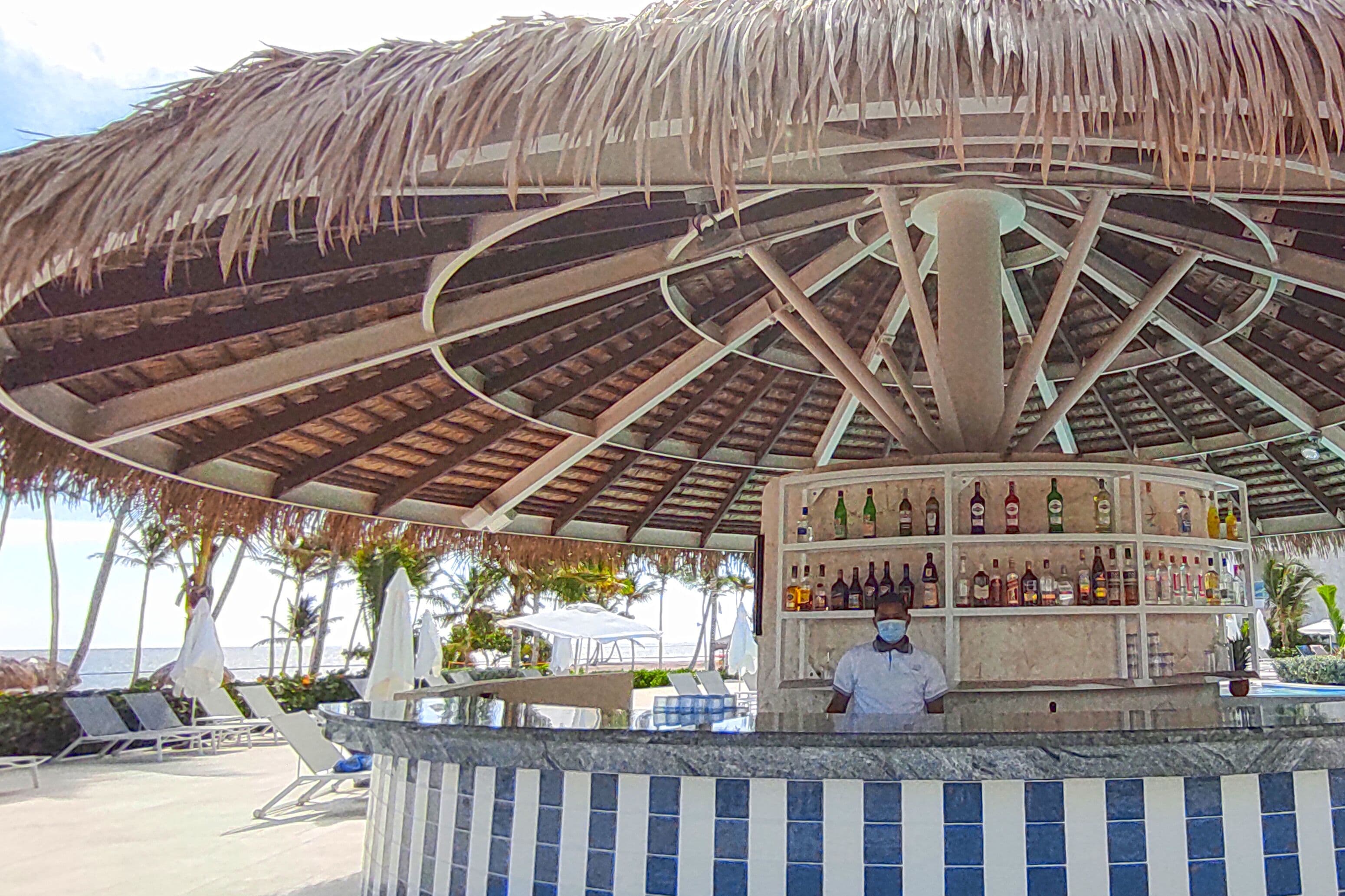Un bar frente al mar con techo de paja, un cantinero y una barra con diversas botellas.
