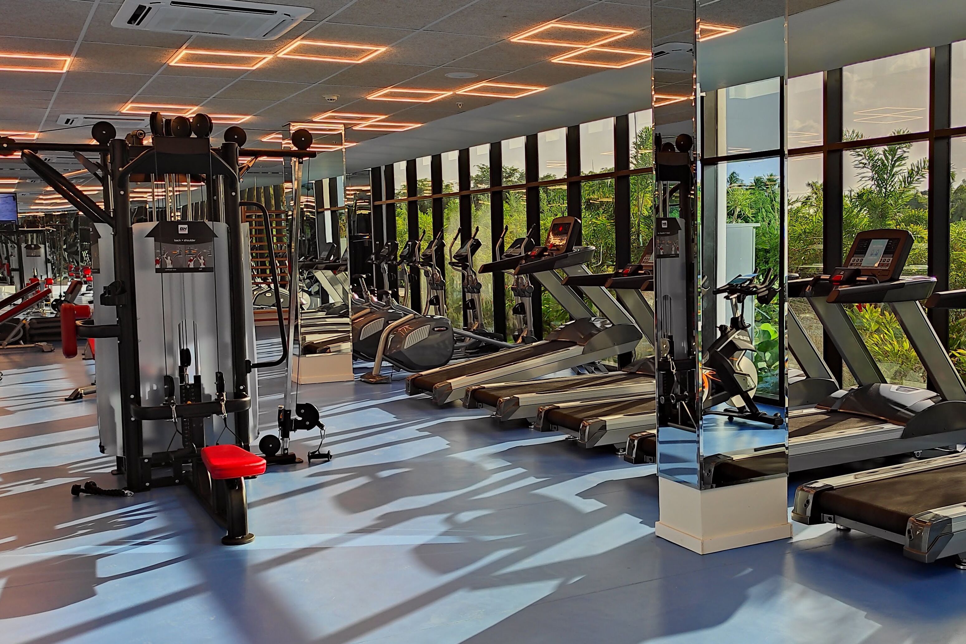 Une salle de sport moderne dotée de divers appareils d’exercice, de grandes fenêtres et d’un sol à motifs.
