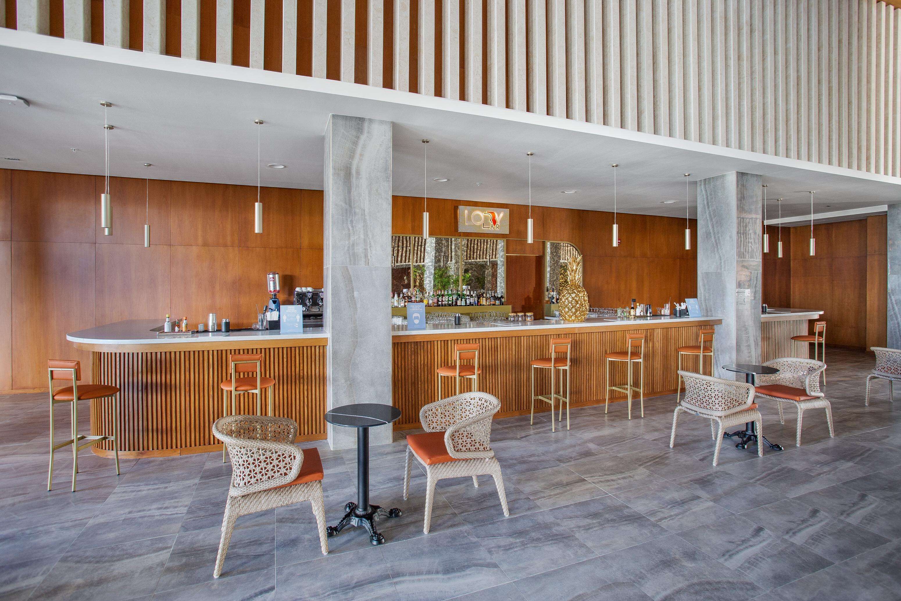 A modern bar area with a wooden bar counter, wicker chairs, and a high ceiling with hanging lights.