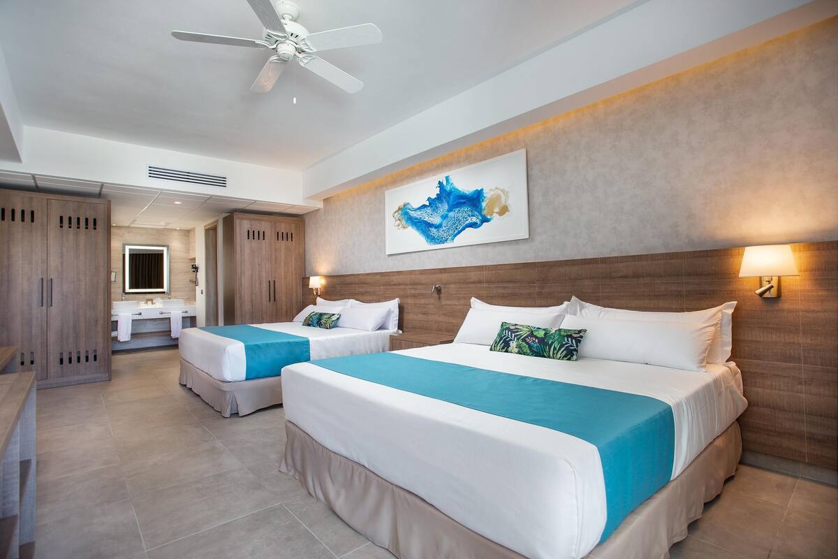 A hotel room with two beds, a ceiling fan, a large artwork on the wall, and a bathroom visible in the background.