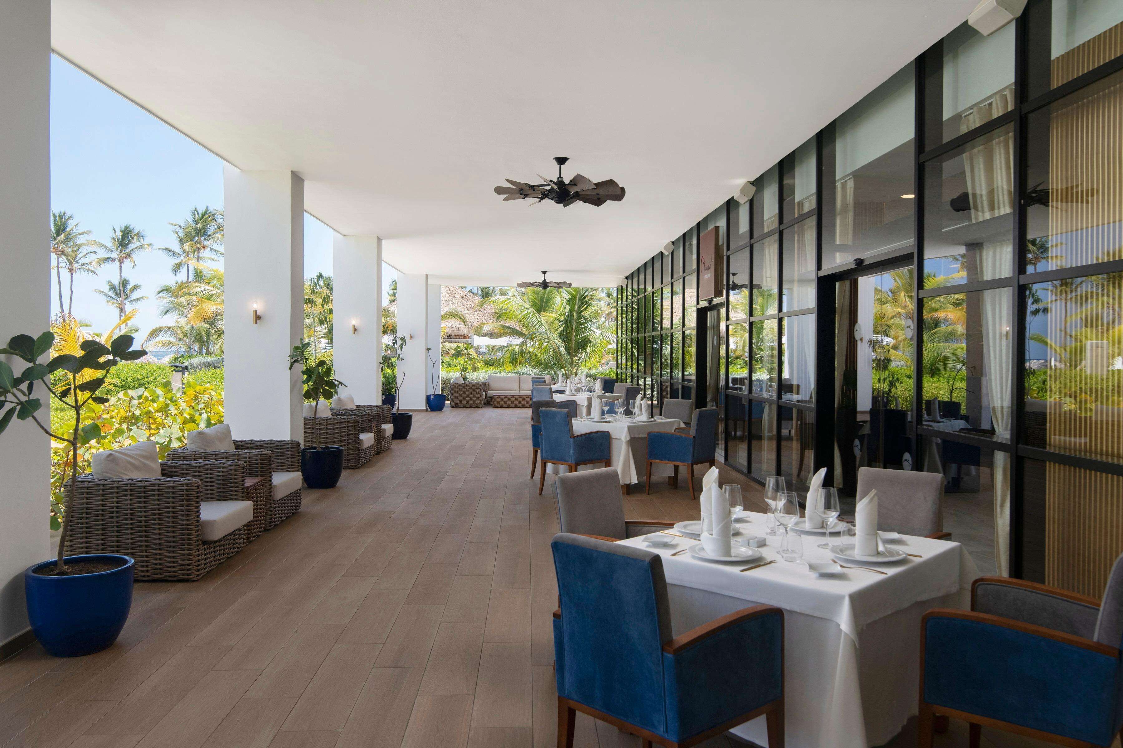 A spacious outdoor dining area with wicker furniture and tables set for dining, large windows offering views of palm trees and greenery, and ceiling fans for ventilation.
