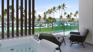 A balcony with a view of palm trees and a pool, furnished with a sofa and a chair. - Serenade Punta Cana Beach & Spa Resort - All Inclusive (Punta Cana)