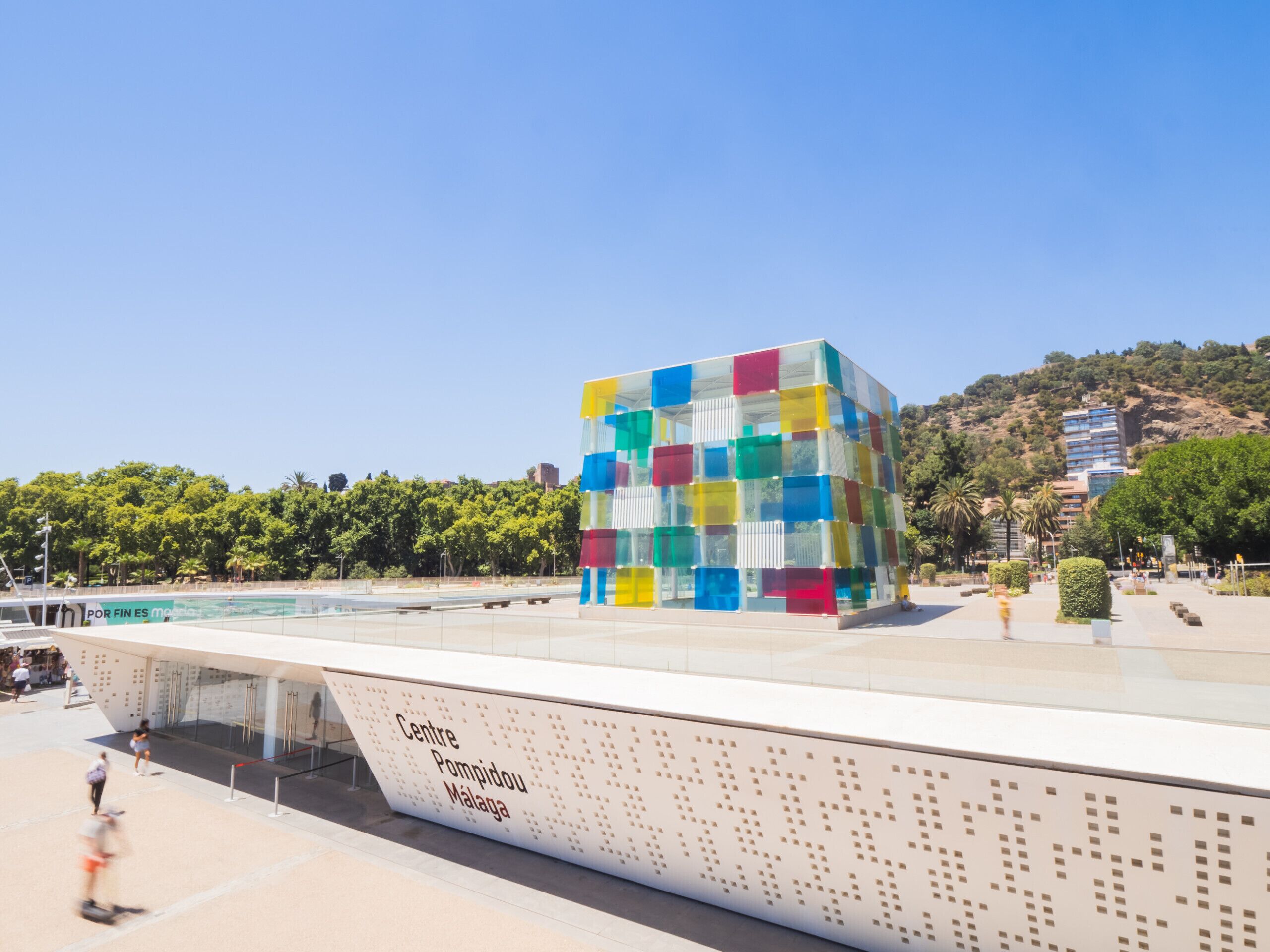 A modern building with a colorful facade, situated in a public square with a clear sky and greenery in the background.