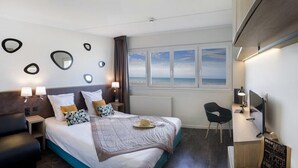 A modern hotel room with a bed, bedside tables, a desk, and a chair. There is a window with a view of the sea.