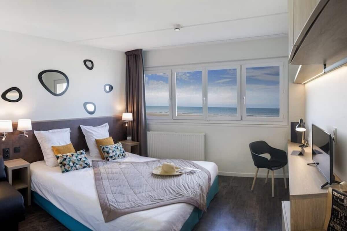 Une chambre d’hôtel moderne équipée d’un lit, de tables de chevet, d’un bureau et d’une chaise. Une fenêtre offre une vue sur la mer.