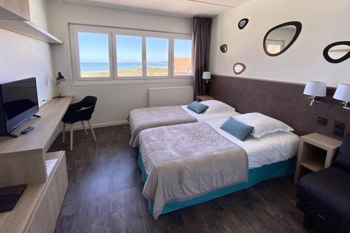 Une chambre d’hôtel avec deux lits, un bureau avec une télévision et une vue sur la plage.