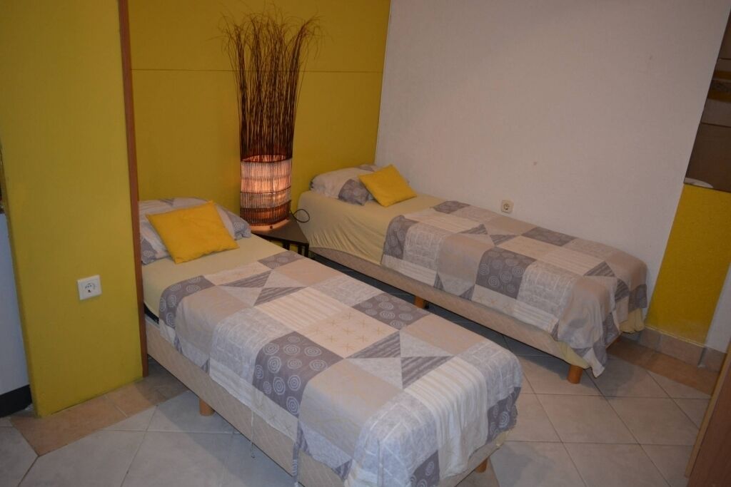 Una stanza con due letti singoli, ciascuno con una coperta a motivi decorativi e un cuscino giallo. Tra i letti è posata un'alta vasi con piante secche.