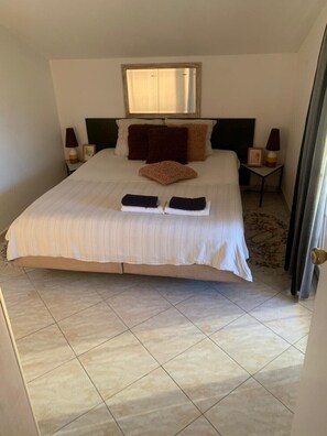 A spacious bedroom with a large bed, two bedside tables with lamps, a headboard, and a window above the bed.