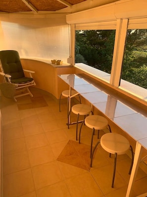 Una habitación con una mesa larga, cuatro taburetes, una silla y un ventanal con vistas a la vegetación.