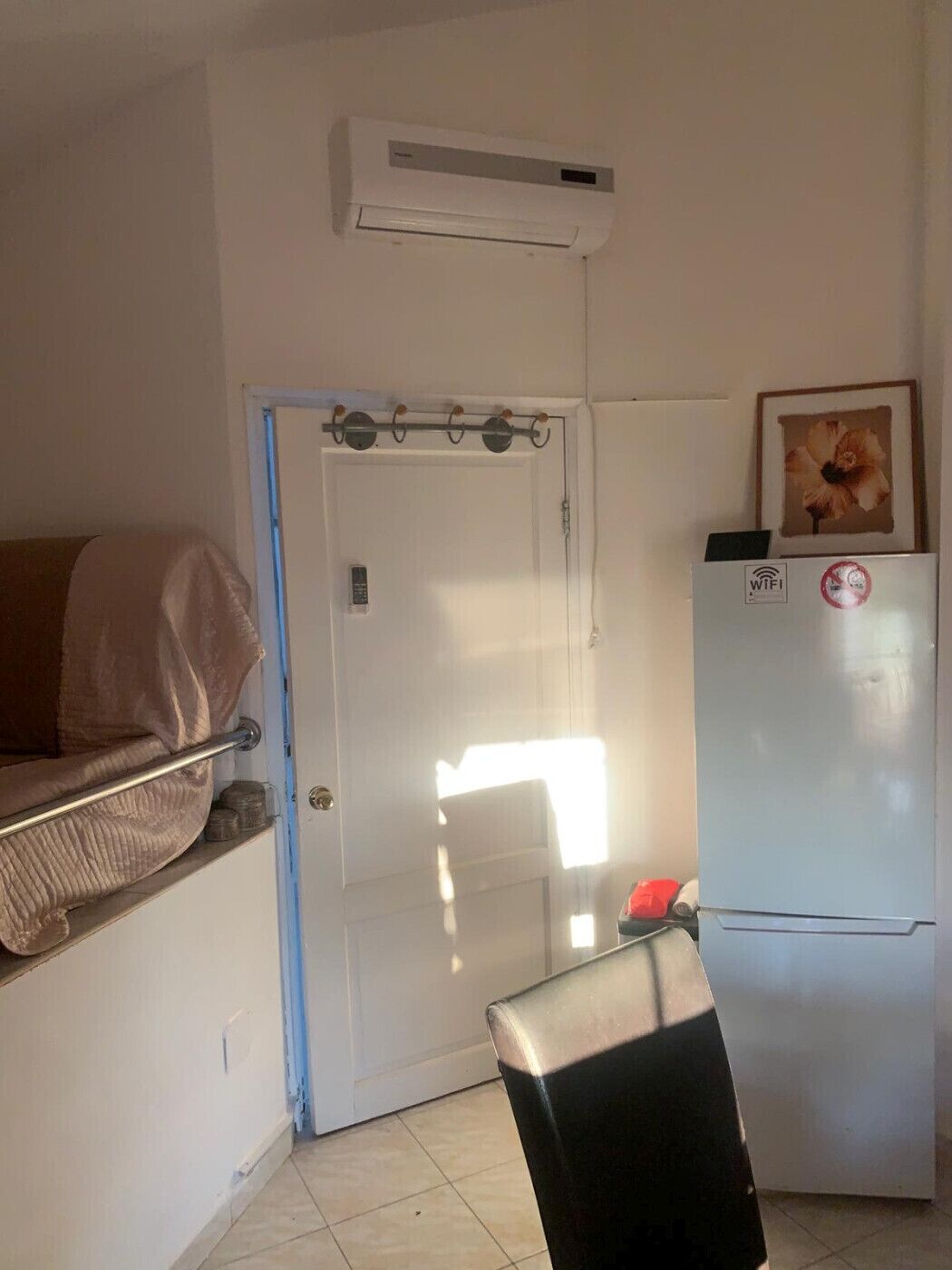 A small room with a white door, a white air conditioner, a white refrigerator, a framed picture on the refrigerator, and a chair in the foreground.