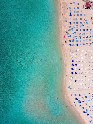 An aerial view of a beach with clear turquoise waters and a sandy shore. There are numerous blue and white sun loungers arranged in rows. - 2 bed Apartment Overlooking Pool - Oroklini (Oroklini)