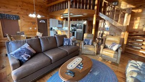 Ein gemütliches Blockhaus-Interieur mit einem braunen Ledersofa, einem runden Couchtisch und einer Treppe, die zu einer oberen Ebene führt.