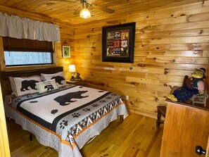 A cozy wooden cabin room with a bed featuring a bear-themed quilt, a small table with a teddy bear, and a framed poster on the wall. - Bear Necessities - Hochatown/ Broken Bow - 3 bd (Broken Bow)