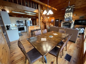 A wooden cabin interior with a dining table, chairs, a kitchen area, and a fireplace. - Bear Necessities - Hochatown/ Broken Bow - 3 bd (Broken Bow)