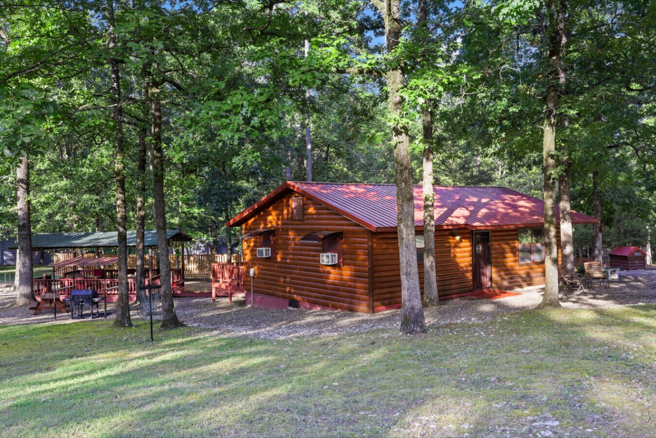 Una cabana rústica amb teulada metàl·lica, envoltada d'alzines altes i una zona de picnic amb taules i cadires.