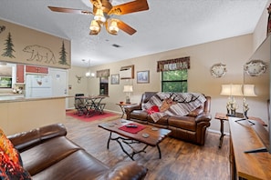 A cozy living room with a brown leather sofa, a wooden coffee table, and a fireplace. There is a dining area with a red rug and a chandelier. The room has a ceiling fan and a window with curtains. - Big Buck Inn- Broken Bow/Hochatown- 2bd (Broken Bow)