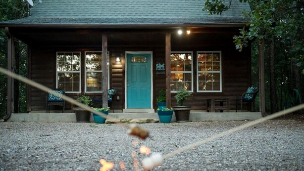 A wooden cabin with a turquoise door, a fire pit in the foreground, and potted plants on the porch.