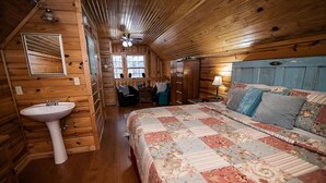 A cozy cabin-style room with a large bed, a wooden floor, a white sink, and a window with curtains.