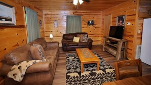 L’intérieur d’un chalet confortable, avec un canapé en cuir, un fauteuil assorti, une table basse en bois et un téléviseur posé sur un meuble en bois.